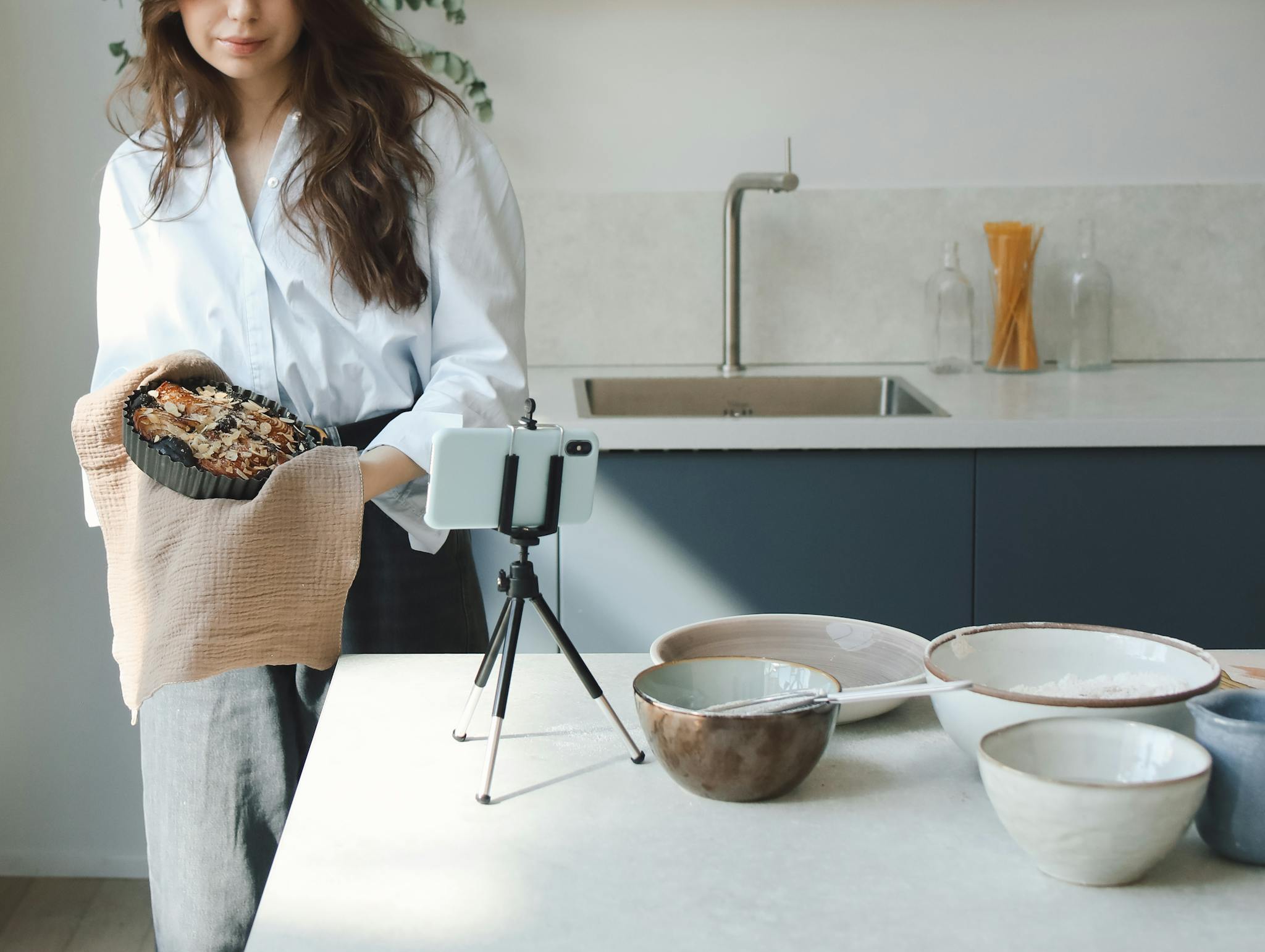 A woman holding a cooked dish in a modern kitchen setting, filming with a smartphone.